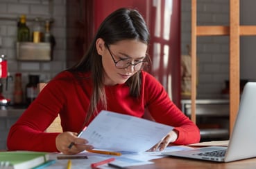 horizontal-shot-project-manager-spectacles-red-juumper-looks-attentively-documents-thinks-how-attract-clients-increase-income-poses-against-kitchen-interior-with-portable-laptop